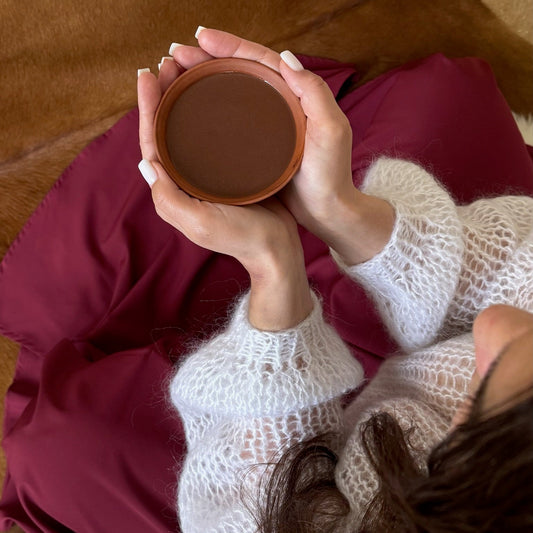 A cozy overhead shot of a person in a white knit sweater and burgundy skirt holding a small brown cup, sitting on a brown leather couch with a flower vase nearby.