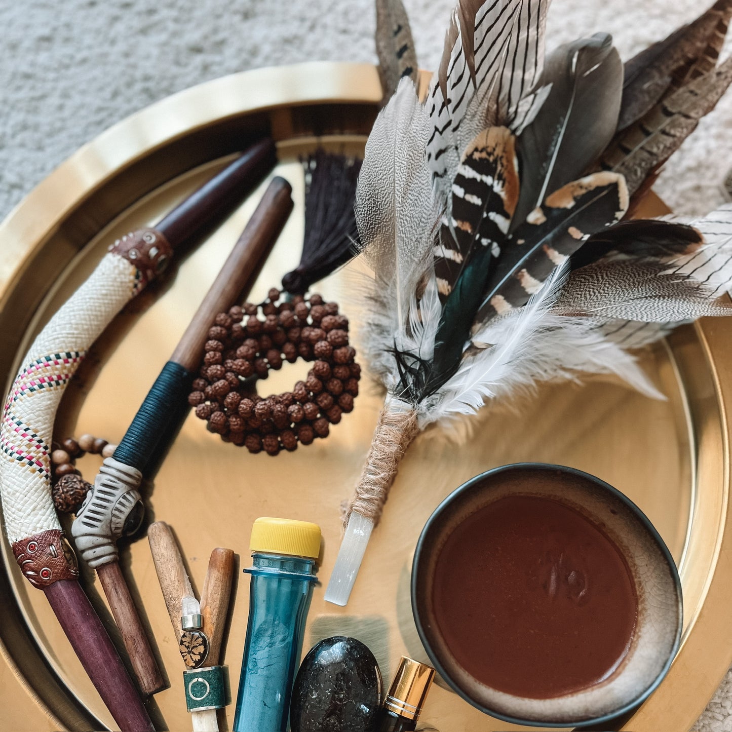 A styled flat-lay of a bohemian self-care and spiritual tray containing a feather wand, wooden beads, a bowl of brown liquid, crystals, and other handmade accessories on a round gold tray.