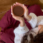 A cozy overhead shot of a person in a white knit sweater and burgundy skirt holding a small brown cup, sitting on a brown leather couch with a flower vase nearby.