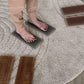 A person wearing beige pants stands barefoot on two gray textured foot mats placed on a round light rug over a wood floor. Two brown massage-like mats are also visible nearby.