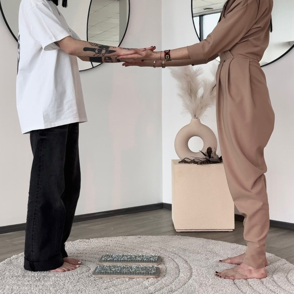 Two women standing barefoot indoors, facing each other and holding hands. One wears a loose white T-shirt and black wide-leg pants, and the other wears a beige wrap-style jumpsuit.